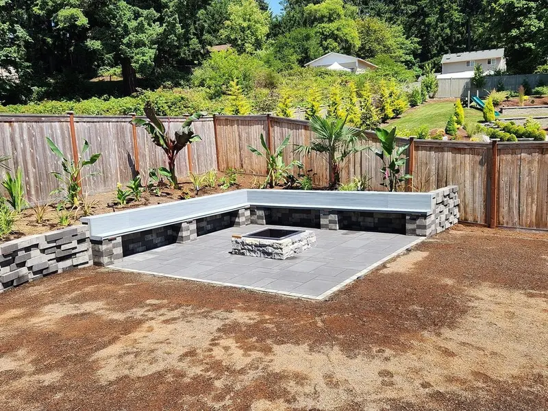 Gray paver patio with a stone fire pit, framed by a built-in block seating wall with light caps and new plantings along a wood fence