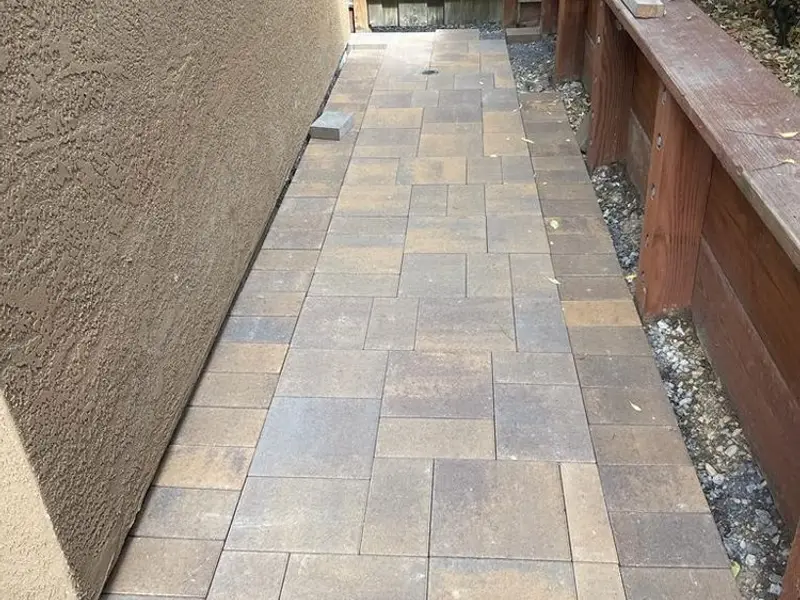 Narrow side-yard walkway of gray pavers along a stucco wall and a wood retaining wall