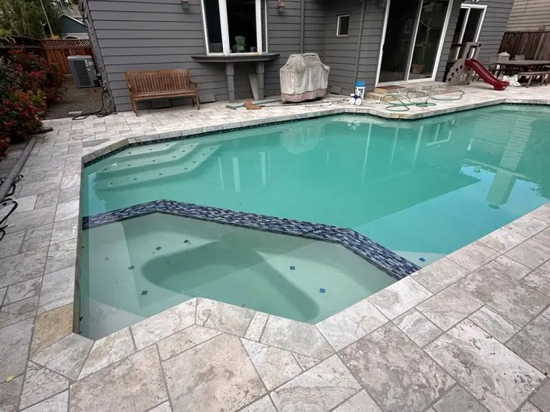 Resurfaced pool with an attached spa, blue mosaic tile along the spa wall, blue step markers and light stone paving around the edge